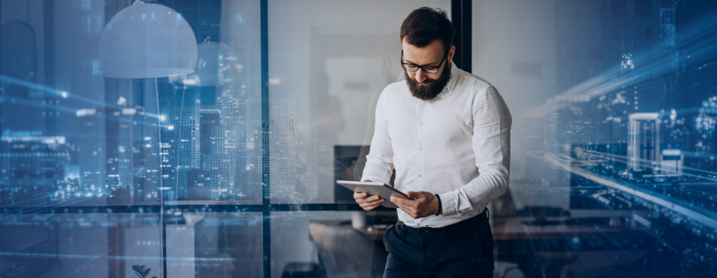 Homem com barba, de camisa branca, olhando para um tablet em um escritório, com uma cidade ao fundo refletida no vidro. Tons de azul criam um efeito tecnológico e omnichannel.