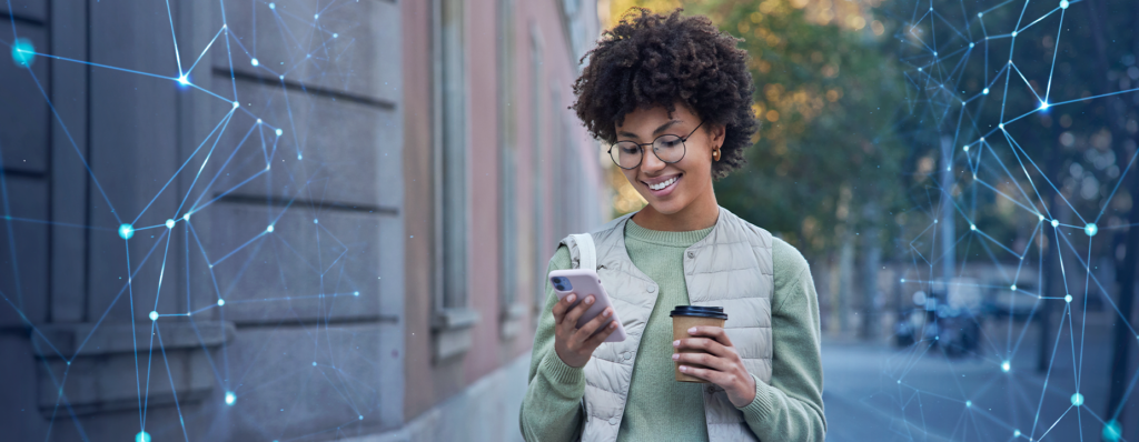 Uma mulher sorridente, com cabelos cacheados e óculos redondos, está olhando para o celular enquanto segura um copo de café. Ela veste uma jaqueta leve e está em uma rua arborizada. Ao redor dela, há uma representação digital de conexões em rede, simbolizando a interatividade e a tecnologia