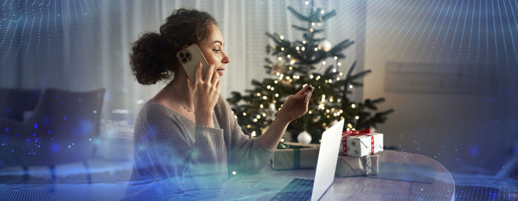 Mulher ao telefone, segurando um cartão de crédito e usando um laptop, com árvore de Natal decorada ao fundo e presentes ao redor.