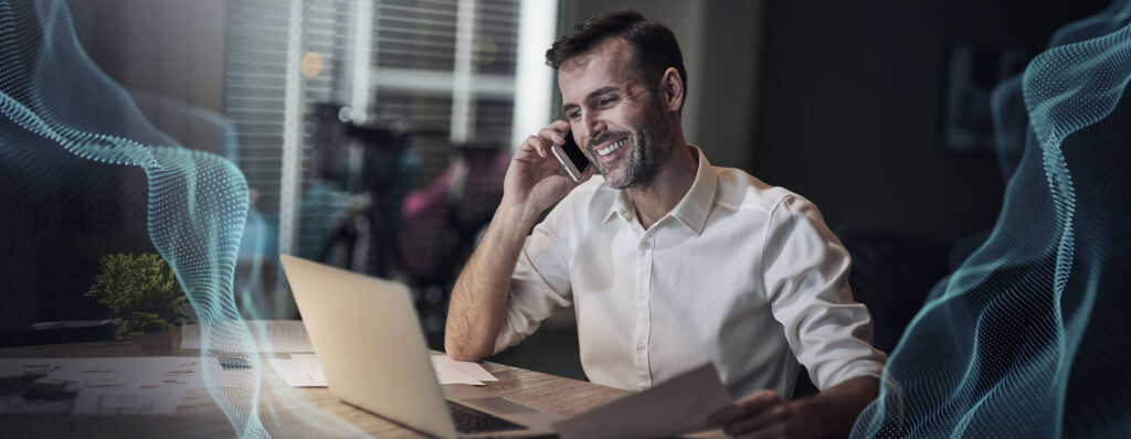 Homem sorridente, usando camisa branca, trabalha em um laptop enquanto fala ao telefone em um ambiente moderno e iluminado, com elementos gráficos digitais ao fundo.