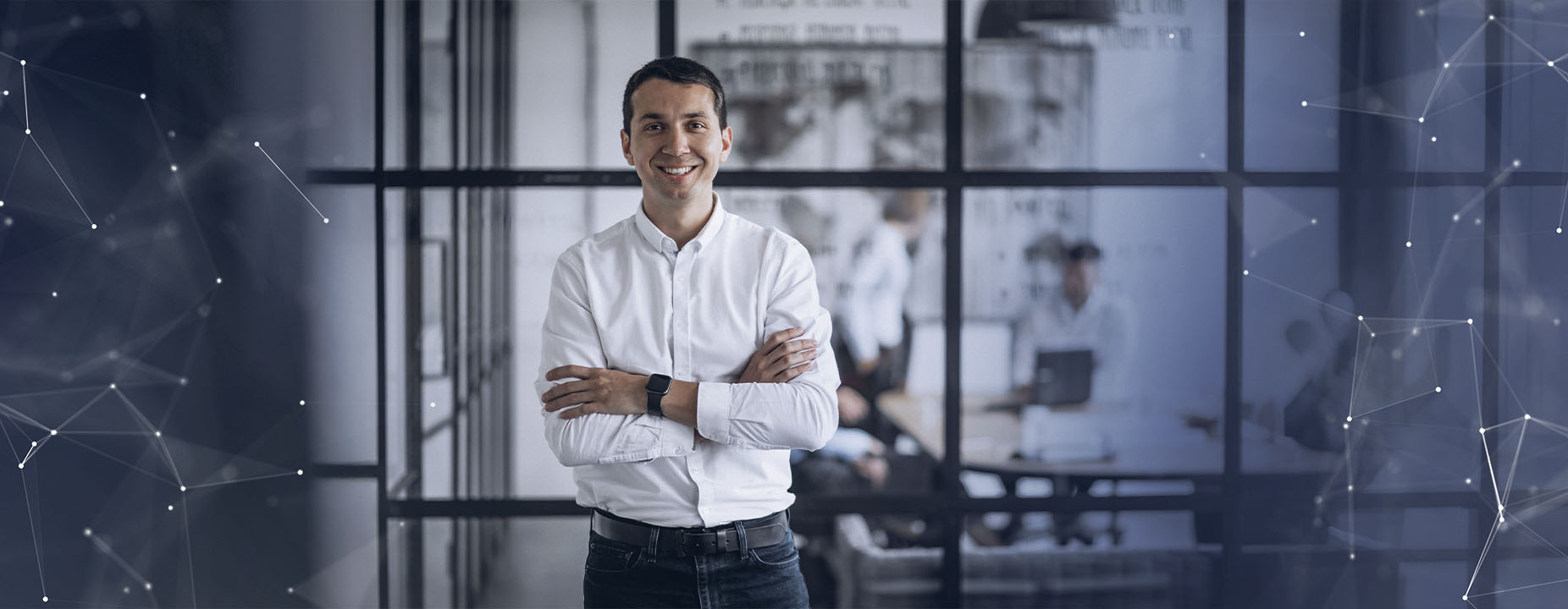 Homem sorridente de braços cruzados, vestindo camisa branca e relógio preto, em ambiente corporativo moderno com paredes de vidro e colegas ao fundo.
